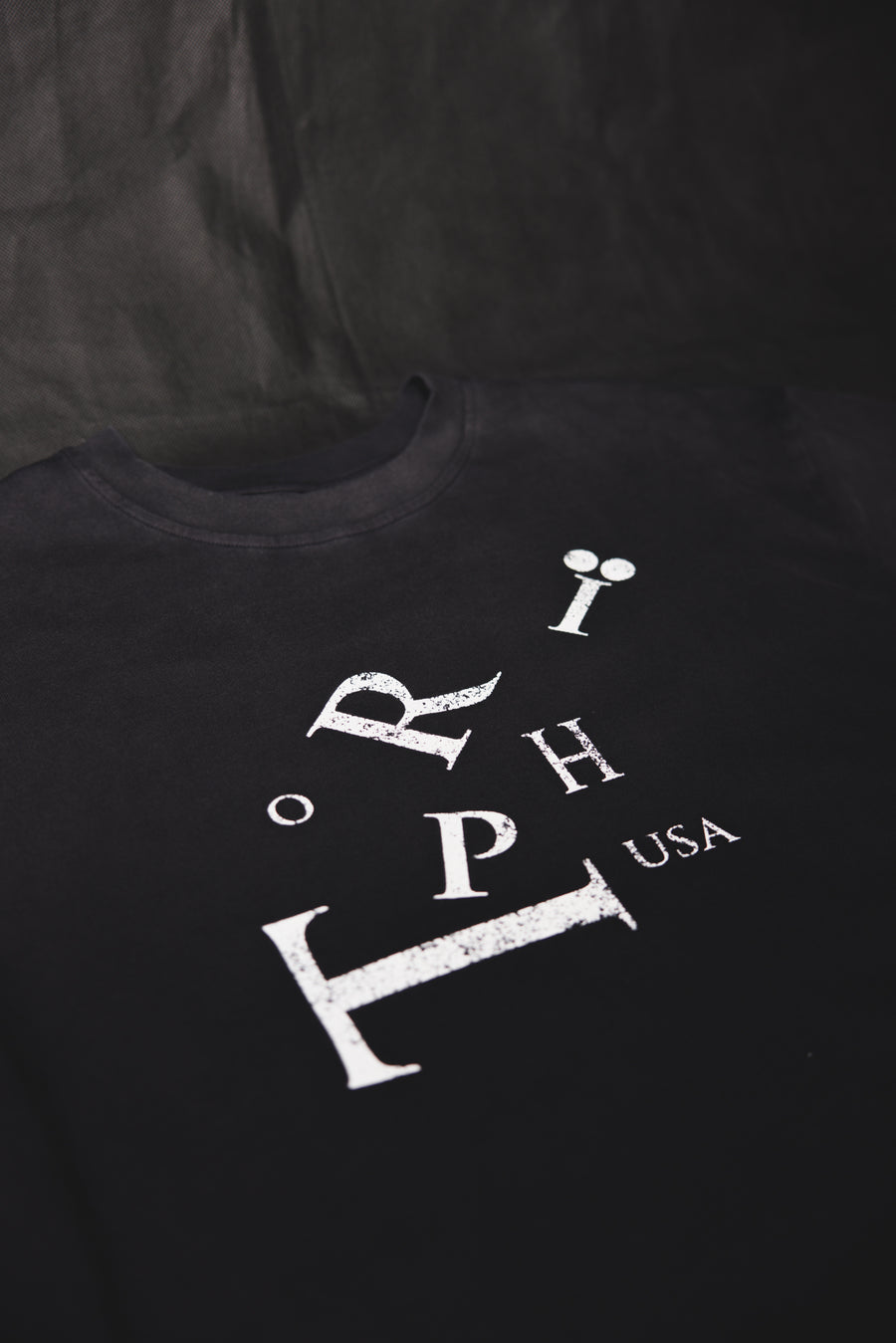 A black crewneck t-shirt featuring a distressed white graphic print of the letters 'RIP' and 'USA' on the front, laid flat against a dark fabric background.