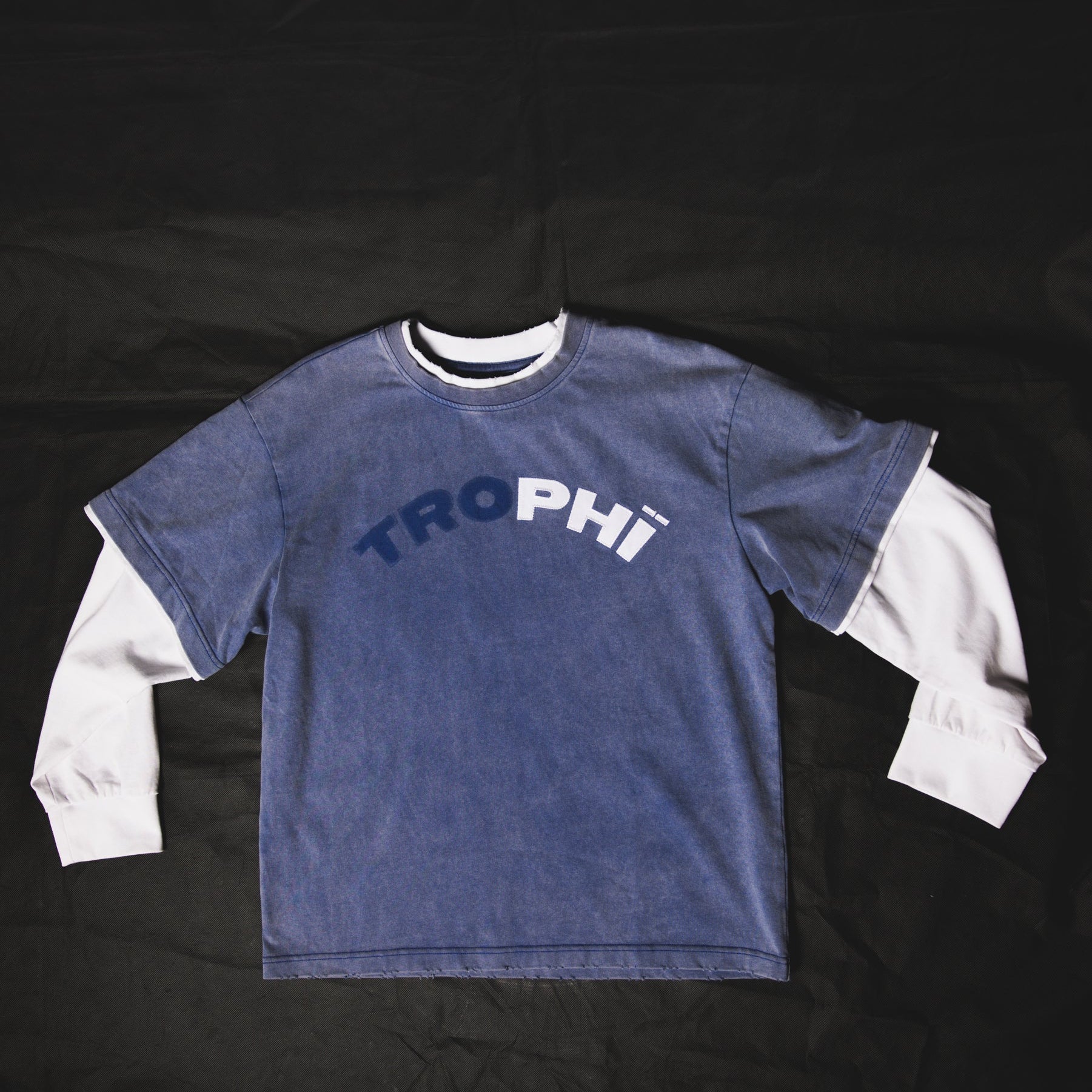 A blue long-sleeve t-shirt with a white layered long-sleeve underneath, laid flat on a black background. The front features the word 'TROPHI' printed in white arched text.